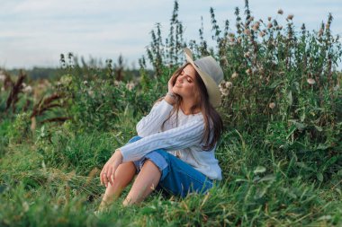 Beyaz kazak, kot pantolon ve kovboy hasır şapkası giyen genç, güzel esmer kadın yeşil çimlerin üzerinde oturuyor, gün batımında gülümsüyor ve gülüyor.