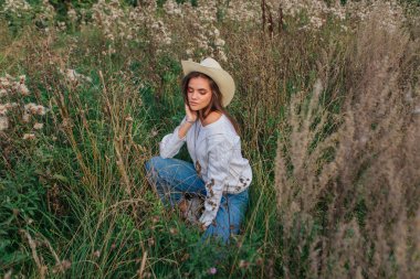 Beyaz kazak, kot pantolon ve kovboy hasır şapkası giyen genç, güzel esmer kadın kuru kahverengi çimlerin üzerinde oturuyor, gün batımında gülümsüyor ve gülüyor.