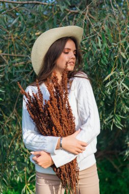 Beyaz kazak, kot pantolon ve kovboy hasır şapkası giymiş genç, güzel esmer kadın elinde kuru yapraklarla ağaç brunch 'larına yakın duruyor.