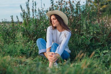 Beyaz kazak, kot pantolon ve kovboy hasır şapkası giyen genç, güzel esmer kadın yeşil çimlerin üzerinde oturuyor, gün batımında gülümsüyor ve gülüyor.