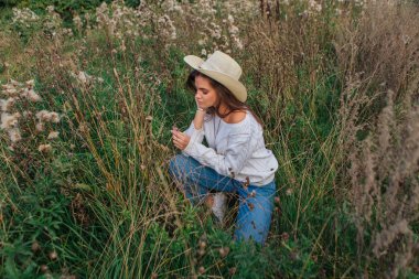 Beyaz kazak, kot pantolon ve kovboy hasır şapkası giyen genç, güzel esmer kadın kuru kahverengi çimlerin üzerinde oturuyor, gün batımında gülümsüyor ve gülüyor.