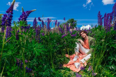 Yeşil ceketli uzun boylu yakışıklı bir adam lupin çiçeklerinin arasında oturmuş, doğanın güzelliğini emrediyor. Mor ve pembe lupinlerle çevrili bir adam..