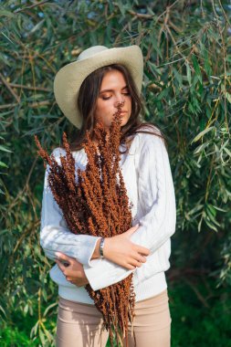 Beyaz kazak, kot pantolon ve kovboy hasır şapkası giymiş genç, güzel esmer kadın elinde kuru yapraklarla ağaç brunch 'larına yakın duruyor.
