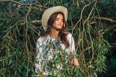 Beyaz kazak, kot pantolon ve kovboy hasır şapkası giymiş genç, güzel esmer kadın yeşil yapraklı ağaç brunch 'larına yakın duruyor.