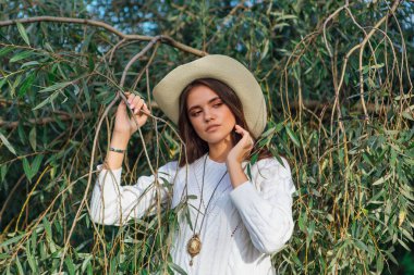 Beyaz kazak, kot pantolon ve kovboy hasır şapkası giymiş genç, güzel esmer kadın yeşil yapraklı ağaç brunch 'larına yakın duruyor.