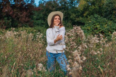 Beyaz kazak, kot pantolon ve kovboy hasır şapkası giymiş genç, güzel esmer kadın kuru kahverengi çimlerin üzerinde duruyor, gün batımında gülümsüyor ve gülüyor.