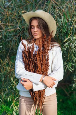 Beyaz kazak, kot pantolon ve kovboy hasır şapkası giymiş genç, güzel esmer kadın elinde kuru yapraklarla ağaç brunch 'larına yakın duruyor.