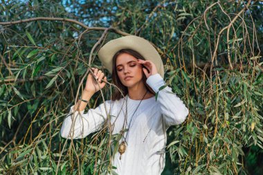 Beyaz kazak, kot pantolon ve kovboy hasır şapkası giymiş genç, güzel esmer kadın yeşil yapraklı ağaç brunch 'larına yakın duruyor.