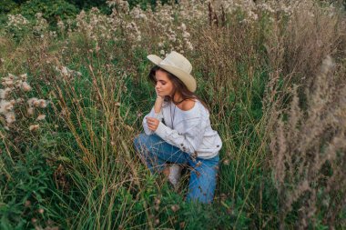 Beyaz kazak, kot pantolon ve kovboy hasır şapkası giyen genç, güzel esmer kadın kuru kahverengi çimlerin üzerinde oturuyor, gün batımında gülümsüyor ve gülüyor.
