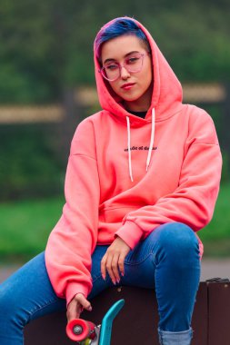 Pembe kapüşonlu ve kot pantolonlu güzel bir kızın portresi kaykayın yanında mavi plastik kaykayıyla oturuyor..