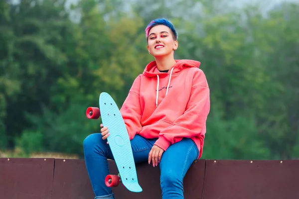 ピンクのパーカーとジーンズに身を包んだピアスの短い色の髪と鼻を持つトレンディーなかなり若い女の子の夏のライフスタイルのイメージ彼女の青いプラスチックスケートボードとスケートボードのコートの横に座って — ストック写真