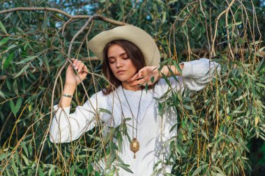 Beyaz kazak, kot pantolon ve kovboy hasır şapkası giymiş genç, güzel esmer kadın yeşil yapraklı ağaç brunch 'larına yakın duruyor.
