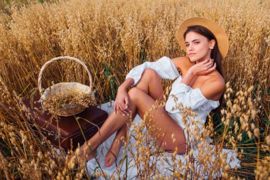 Kırsal Kır Sahnesi. Beyaz bluz ve hasır şapka giymiş uzun saçlı güzel bir kadın altın yulaf tarlasında oturmuş yulaf kulaklı sepeti tutuyor..