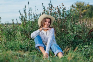 Beyaz kazak, kot pantolon ve kovboy hasır şapkası giyen genç, güzel esmer kadın yeşil çimlerin üzerinde oturuyor, gün batımında gülümsüyor ve gülüyor.