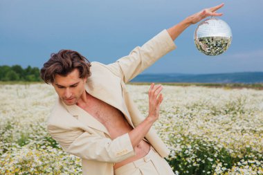 Beyaz takım elbiseli uzun boylu, çıplak bir adam papatya çiçeği tarlasında dikiliyor, elinde ayna disko topu tutuyor..