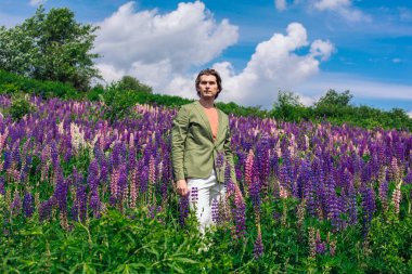 Yeşil ceketli uzun boylu yakışıklı bir adam lupin çiçeği tarlasında duruyor, doğanın güzelliğini emrediyor. Mor ve pembe lupinlerle çevrili bir adam..