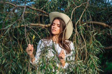 Genç ve güzel esmer kadın ağaç brançlarının yanında yeşil yapraklarla duruyor.