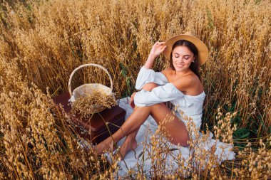 Hasır şapkalı genç bir kadın altın yulaf tarlasında oturmuş yulaf kulaklı sepeti tutuyor..
