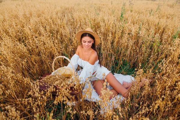 Joven hermosa mujer con sombrero de paja en la cabeza que pone en el campo de avena de oro que sostiene la cesta con orejas de avena. — Foto de Stock