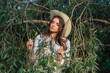 Beyaz kazak, kot pantolon ve kovboy hasır şapkası giymiş genç, güzel esmer kadın yeşil yapraklı ağaç brunch 'larına yakın duruyor.