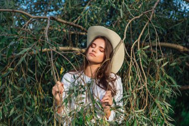 Beyaz kazak, kot pantolon ve kovboy hasır şapkası giymiş genç, güzel esmer kadın yeşil yapraklı ağaç brunch 'larına yakın duruyor.