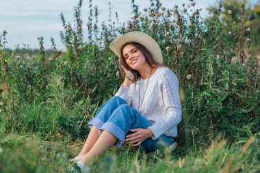 Beyaz kazak, kot pantolon ve kovboy hasır şapkası giyen genç, güzel esmer kadın yeşil çimlerin üzerinde oturuyor, gün batımında gülümsüyor ve gülüyor.