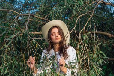 Beyaz kazak, kot pantolon ve kovboy hasır şapkası giymiş genç, güzel esmer kadın yeşil yapraklı ağaç brunch 'larına yakın duruyor.