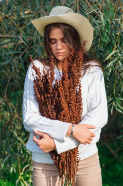 Beyaz kazak, kot pantolon ve kovboy hasır şapkası giymiş genç, güzel esmer kadın elinde kuru yapraklarla ağaç brunch 'larına yakın duruyor.