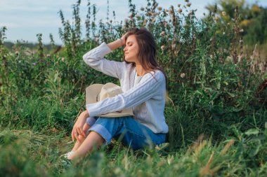 Beyaz kazak, kot pantolon ve kovboy hasır şapkası giyen genç, güzel esmer kadın yeşil çimlerin üzerinde oturuyor, gün batımında gülümsüyor ve gülüyor.