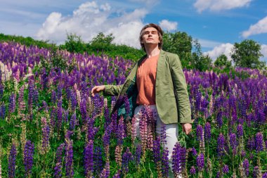 Yeşil ceketli uzun boylu yakışıklı bir adam lupin çiçeği tarlasında dans ediyor, doğanın güzelliğini emrediyor. Mor ve pembe lupinlerle çevrili bir adam..