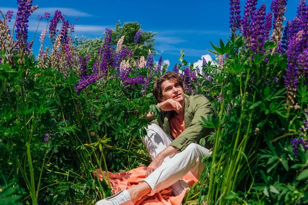 Tall handsome man in a green jacket sitting among the lupine flowers, enjoing the beauty of nature. Man surrounded by purple and pink lupines.