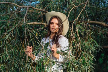 Beyaz kazak, kot pantolon ve kovboy hasır şapkası giymiş genç, güzel esmer kadın yeşil yapraklı ağaç brunch 'larına yakın duruyor.