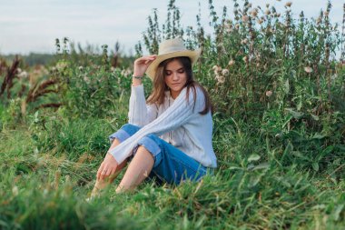 Beyaz kazak, kot pantolon ve kovboy hasır şapkası giyen genç, güzel esmer kadın yeşil çimlerin üzerinde oturuyor, gün batımında gülümsüyor ve gülüyor.