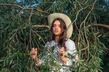 Beyaz kazak, kot pantolon ve kovboy hasır şapkası giymiş genç, güzel esmer kadın yeşil yapraklı ağaç brunch 'larına yakın duruyor.