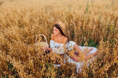 Kırsal Kır Sahnesi. Beyaz bluz ve hasır şapka giymiş, uzun saçlı genç ve güzel bir kadın altın yulaf tarlasında, yulaf kulaklı sepeti tutuyordu..