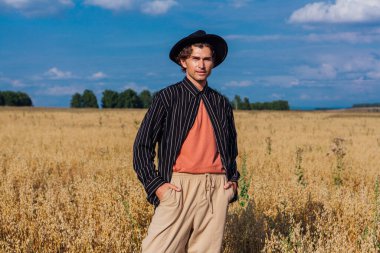 Kırsal Kır Sahnesi. Siyah gömlekli ve siyah şapkalı uzun boylu yakışıklı adam altın yulaf tarlasında duruyordu. Mavi gökyüzü ile yaz manzarası