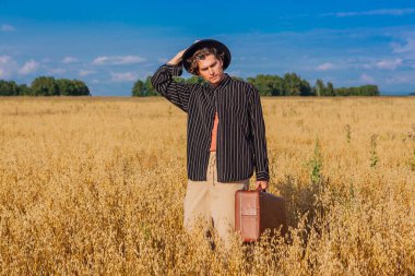 Kırsal Kır Sahnesi. Siyah gömlekli ve siyah şapkalı uzun boylu yakışıklı adam altın yulaf tarlasında kahverengi deri valiziyle dikiliyordu. Mavi gökyüzü ile yaz manzarası
