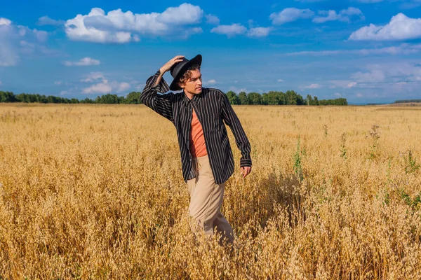 Rural Countryside Scene. Tall handsome man dressed in a black shirt and black hat standing at golden oat field. Summer landscape with blue sky