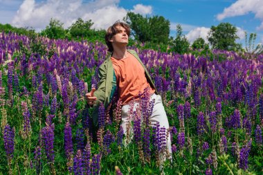 Yeşil ceketli uzun boylu yakışıklı bir adam lupin çiçeği tarlasında dans ediyor, doğanın güzelliğini emrediyor. Mor ve pembe lupinlerle çevrili bir adam..