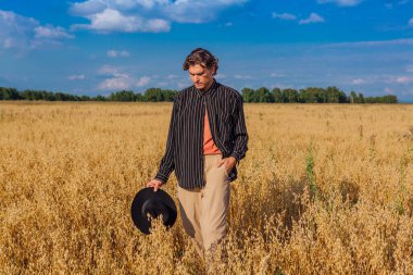 Kırsal Kır Sahnesi. Siyah gömlekli ve siyah şapkalı uzun boylu yakışıklı adam altın yulaf tarlasında duruyordu. Mavi gökyüzü ile yaz manzarası