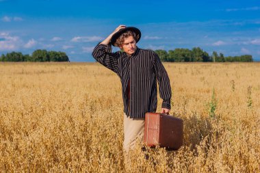 Kırsal Kır Sahnesi. Siyah gömlekli ve siyah şapkalı uzun boylu yakışıklı adam altın yulaf tarlasında kahverengi deri valiziyle dikiliyordu. Mavi gökyüzü ile yaz manzarası