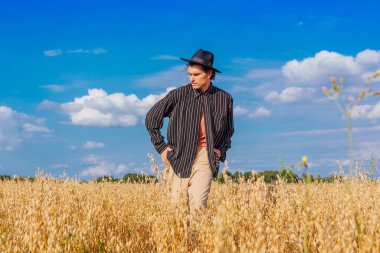 Kırsal Kır Sahnesi. Siyah gömlekli ve siyah şapkalı uzun boylu yakışıklı adam altın yulaf tarlasında duruyordu. Mavi gökyüzü ile yaz manzarası