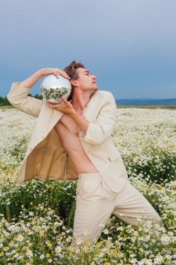 Beyaz takım elbiseli uzun boylu, çıplak bir adam papatya çiçeği tarlasında dikiliyor, elinde ayna disko topu tutuyor..