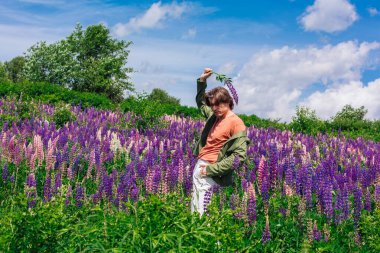Yeşil ceketli uzun boylu yakışıklı bir adam lupin çiçeği tarlasında duruyor elinde lupin çiçekleri, doğanın güzelliğini emrediyor. Mor ve pembe lupinlerle çevrili bir adam..