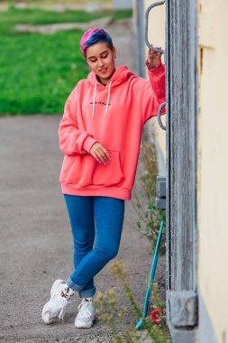 Kısa saçlı, pembe kapüşonlu, eski tahta kapıların yanında plastik kaykayıyla duran zarif bir genç kadın. Gençlik kavramı.