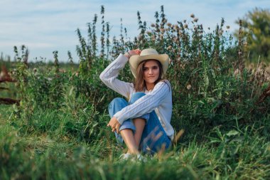 Beyaz kazak, kot pantolon ve kovboy hasır şapkası giyen genç, güzel esmer kadın yeşil çimlerin üzerinde oturuyor, gün batımında gülümsüyor ve gülüyor.