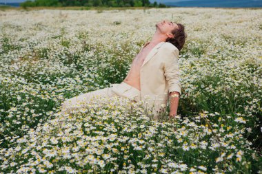 Beyaz takım elbiseli uzun boylu yakışıklı adam papatya çiçeği tarlasında beyaz bir sandalyede oturuyor.