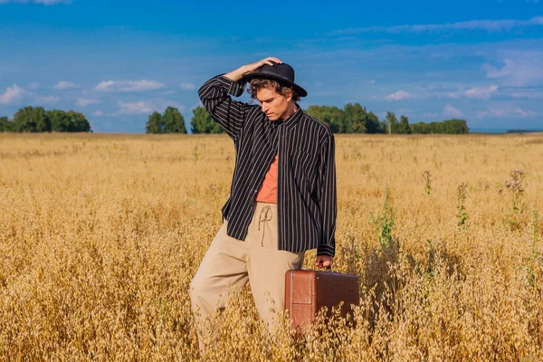 Rural Countryside Scene. Tall handsome man dressed in a black shirt and black hat standing at golden oat field with brown vintage leather suitcase. Summer landscape with blue sky