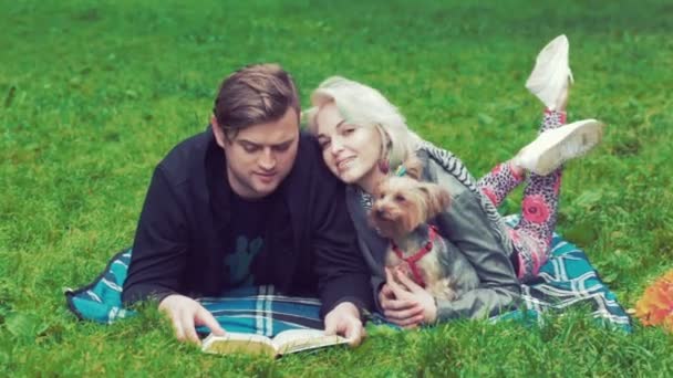 Un disparo de una joven pareja con un perro. Se acuestan en la hierba y discuten lo que leen en el libro — Vídeo de stock