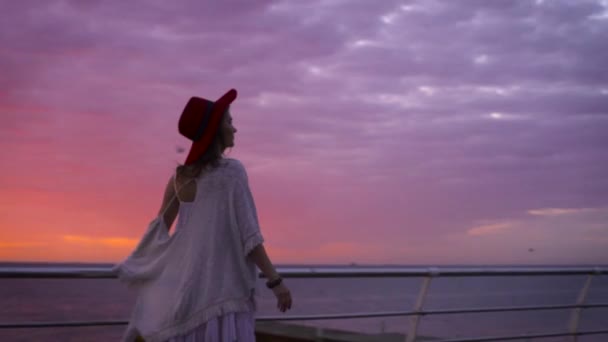 白いドレスとすばらしい夕日で海の背景にポーズの帽子を身に着けている若い魅力的な女性 — ストック動画